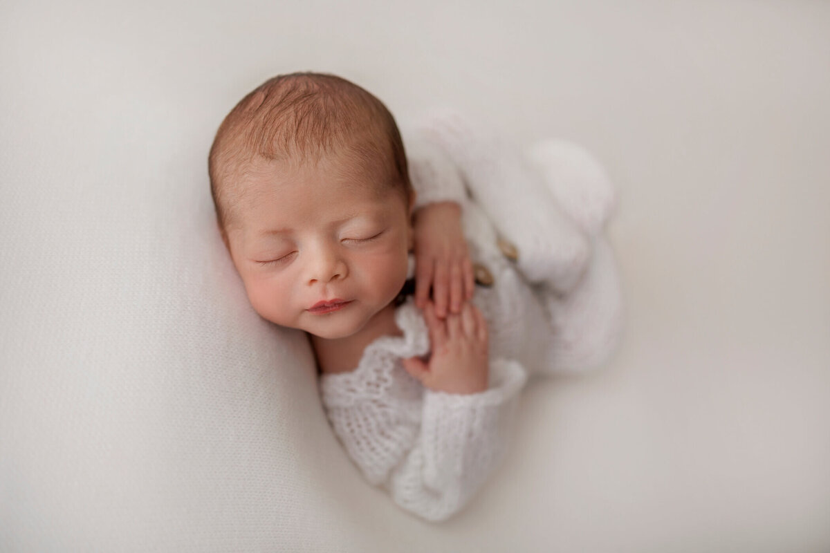 Newborn baby boy peacefully sleeping on a white blanket, dressed in a cozy white knitted outfit. His hands are gently placed on his chest.