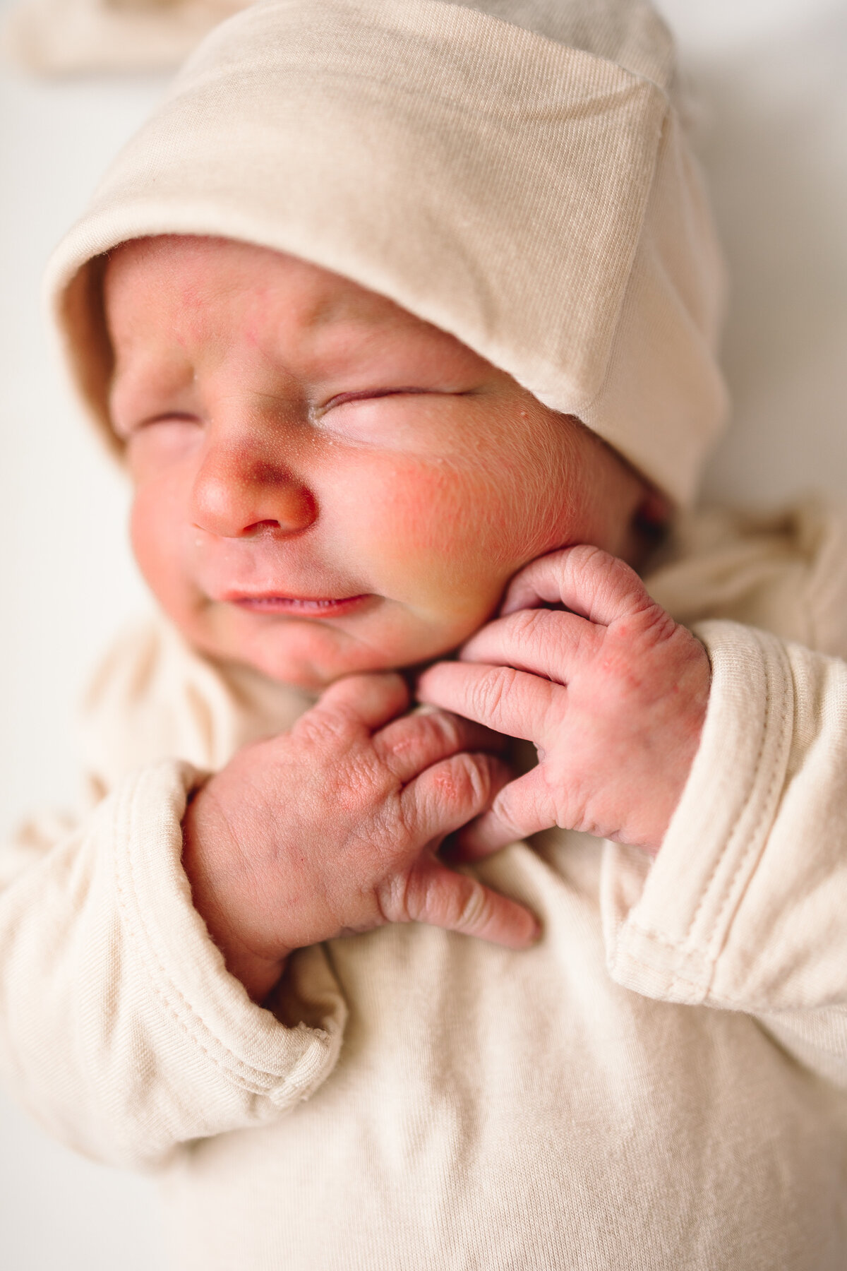 Newborn baby sleeping peacefully on a white blanket. The baby has eyes closed, hands resting on the chest, and is dressed in a cozy outfit with a beige hat. Captured by an Albuquerque lifestyle maternity photographer