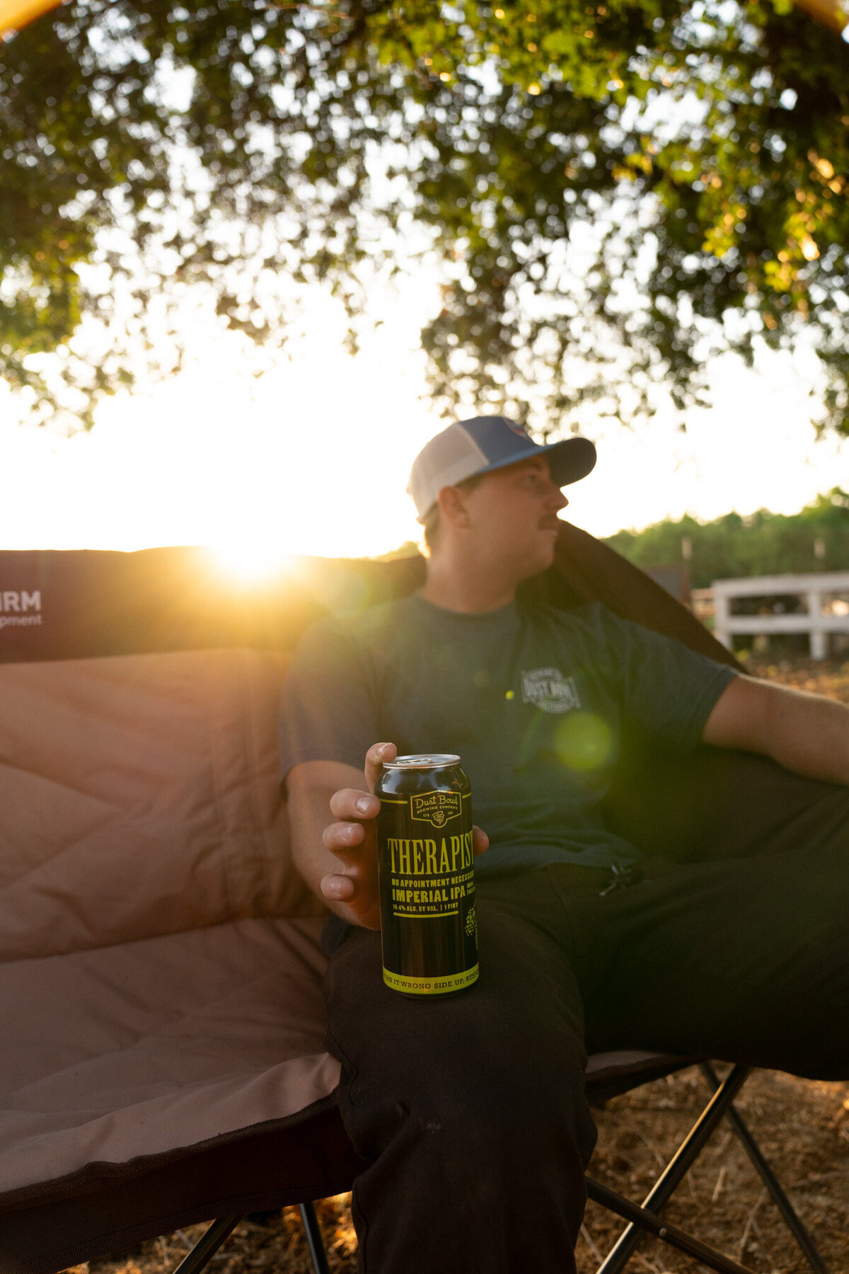 Man wearing a Dust Bowl Brewing hat and tshirt sitting in a lawn chair at sunset with a Therapist IPA in his hand