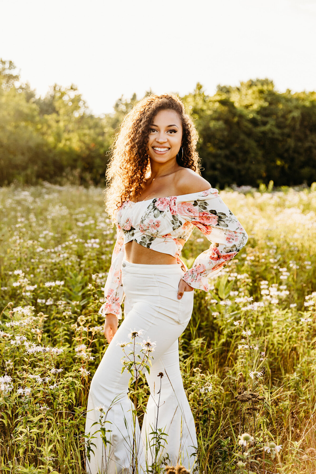 teen in a floral crop top and white flare pants smiling in a field of wildflowers