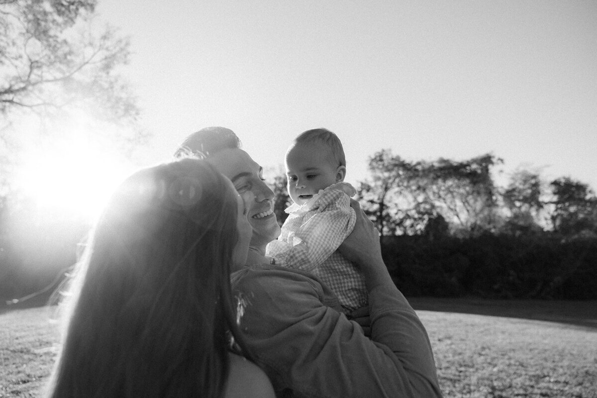 Infant is held by father while mother looks on and family is washed in sunlight. 