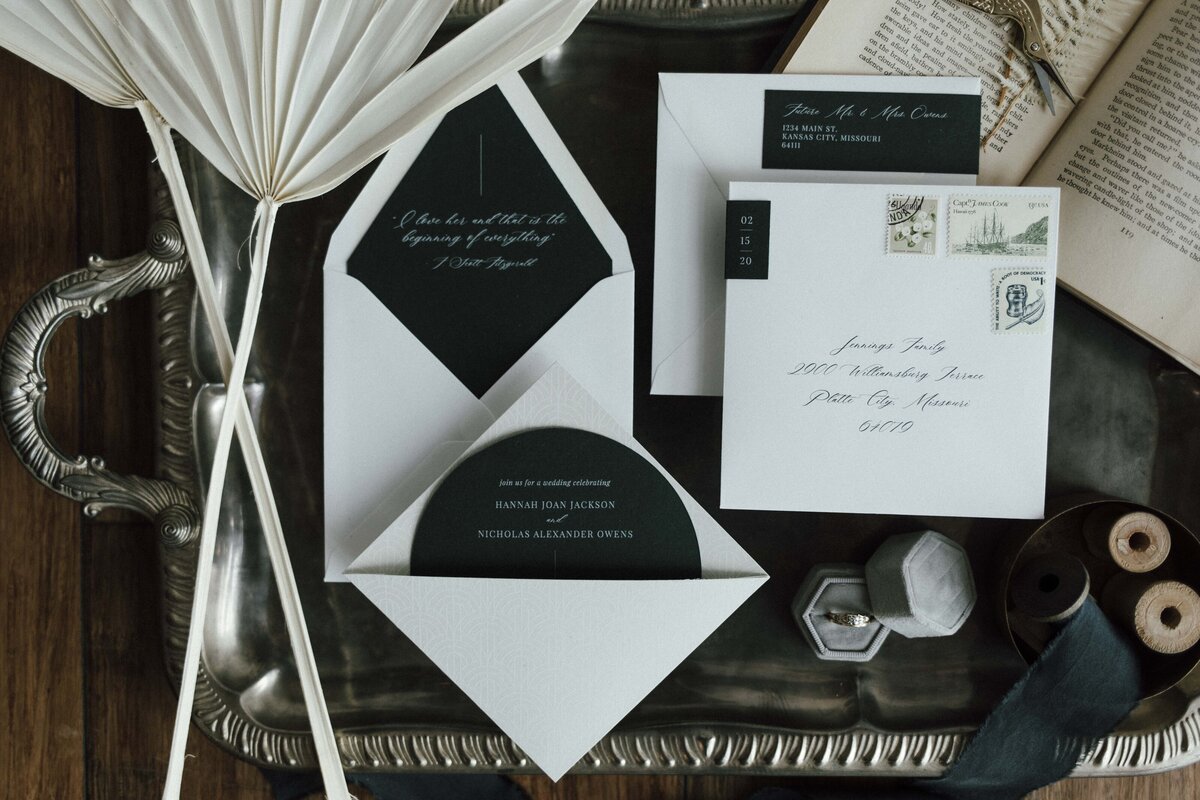 Mixed white wedding stationery with cursive fonts set on a silver tray with a book and scissors on a wooden floor.