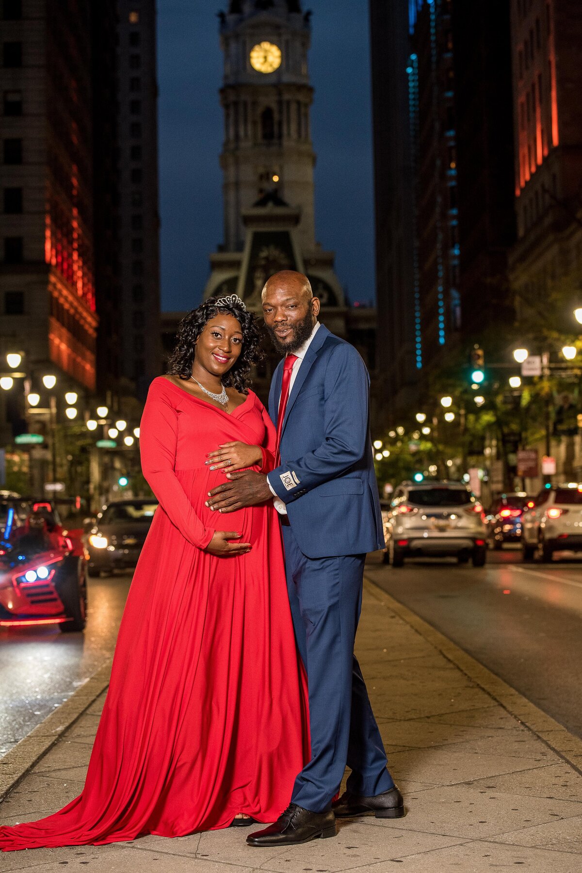 a Black woman and man post in front of Downtown Austin. She wears a red maternity dress and he's wearing a blue suit