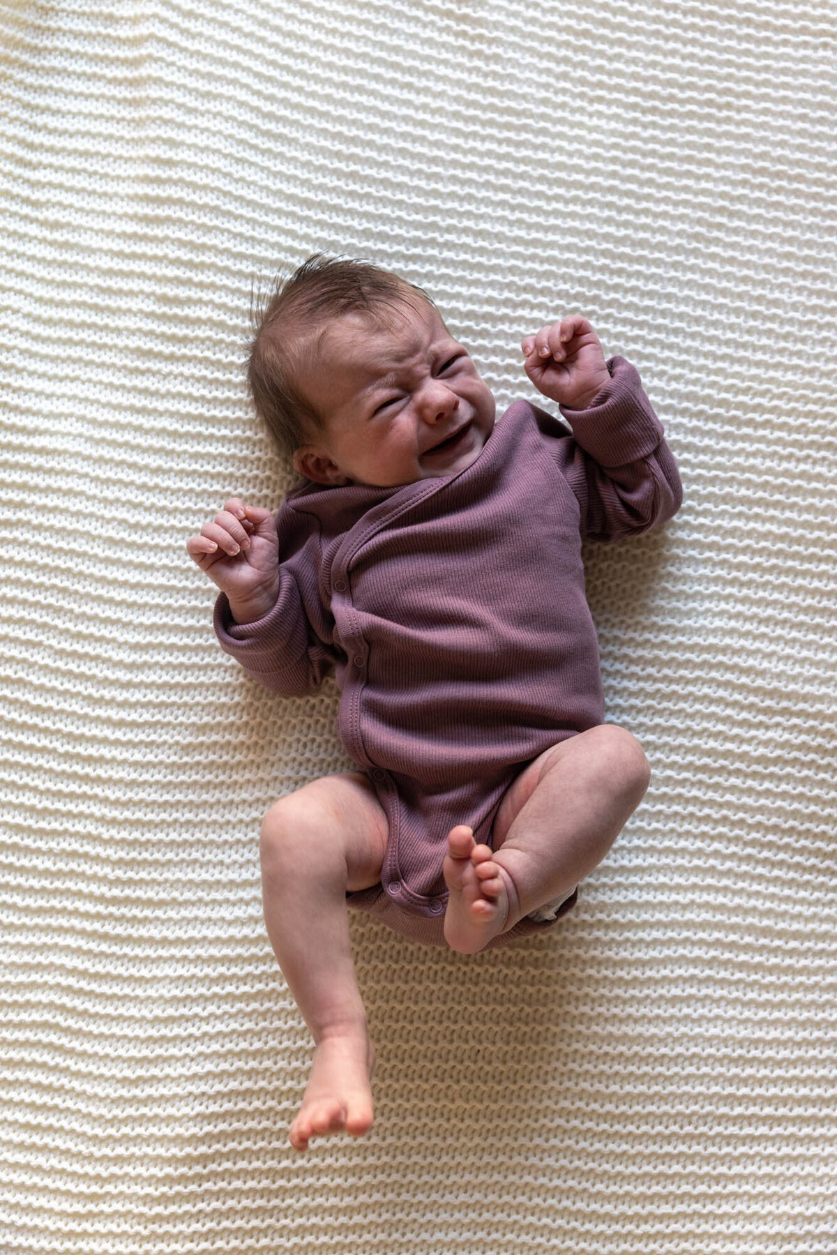 Foto av baby i lilla body som ligger på et strikket hvitt pledd. Babyen gråter og spreller.