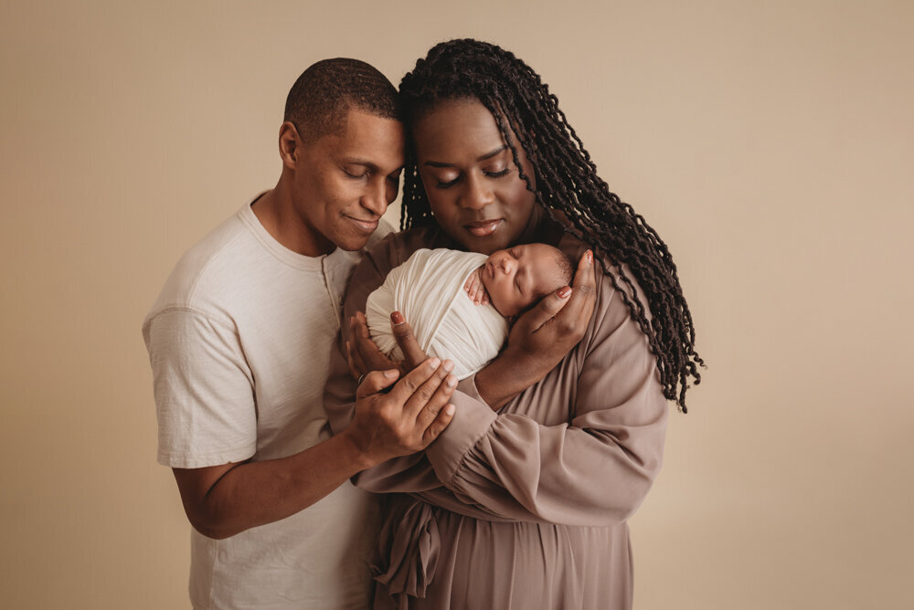 Newborn photos with Atlanta, Georgia newborn photographer of sleeping baby boy with hands under chin wrapped in cream swaddle being held by his  parents.