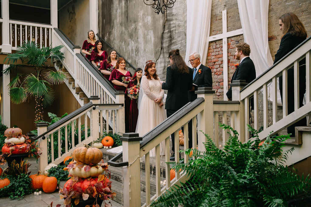A bride and groom stand on a staircase, exchanging vows in an intimate wedding ceremony. Bridesmaids in burgundy dresses and groomsmen in black suits look on. Autumn decorations, pumpkins, and ferns surround the scene.
