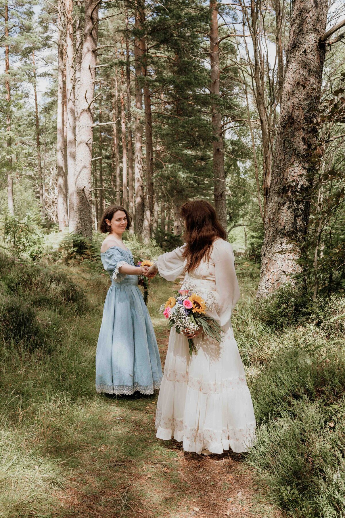 An LGBTQ+ couple in vintage dresses stand in a lush Aberdeenshire forest, posing for their wedding photography portraits. One holds a bouquet of colourful flowers, and they face each other amidst tall trees and greenery, creating a serene, pastoral scene under a partly cloudy sky.
