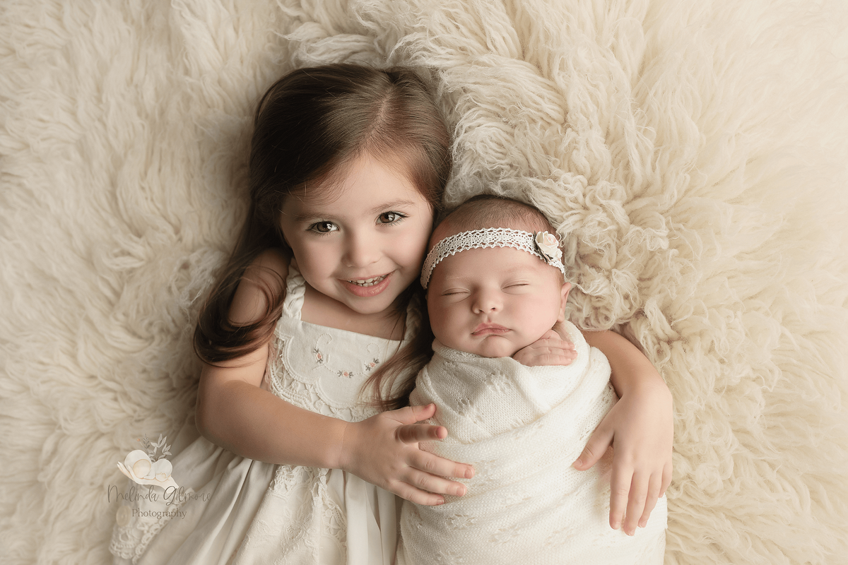 A young girl in a white dress cuddles with her sleeping newborn baby sister on a fur blanket in a studio taken by a New Orleans Newborn Photographer