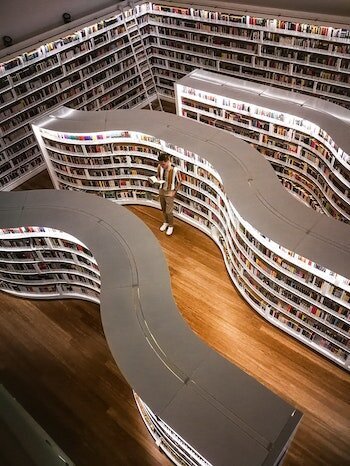 Photo from above of a man reading a book in curving library stacks. Photo by Mark Cruzat via Pexels.