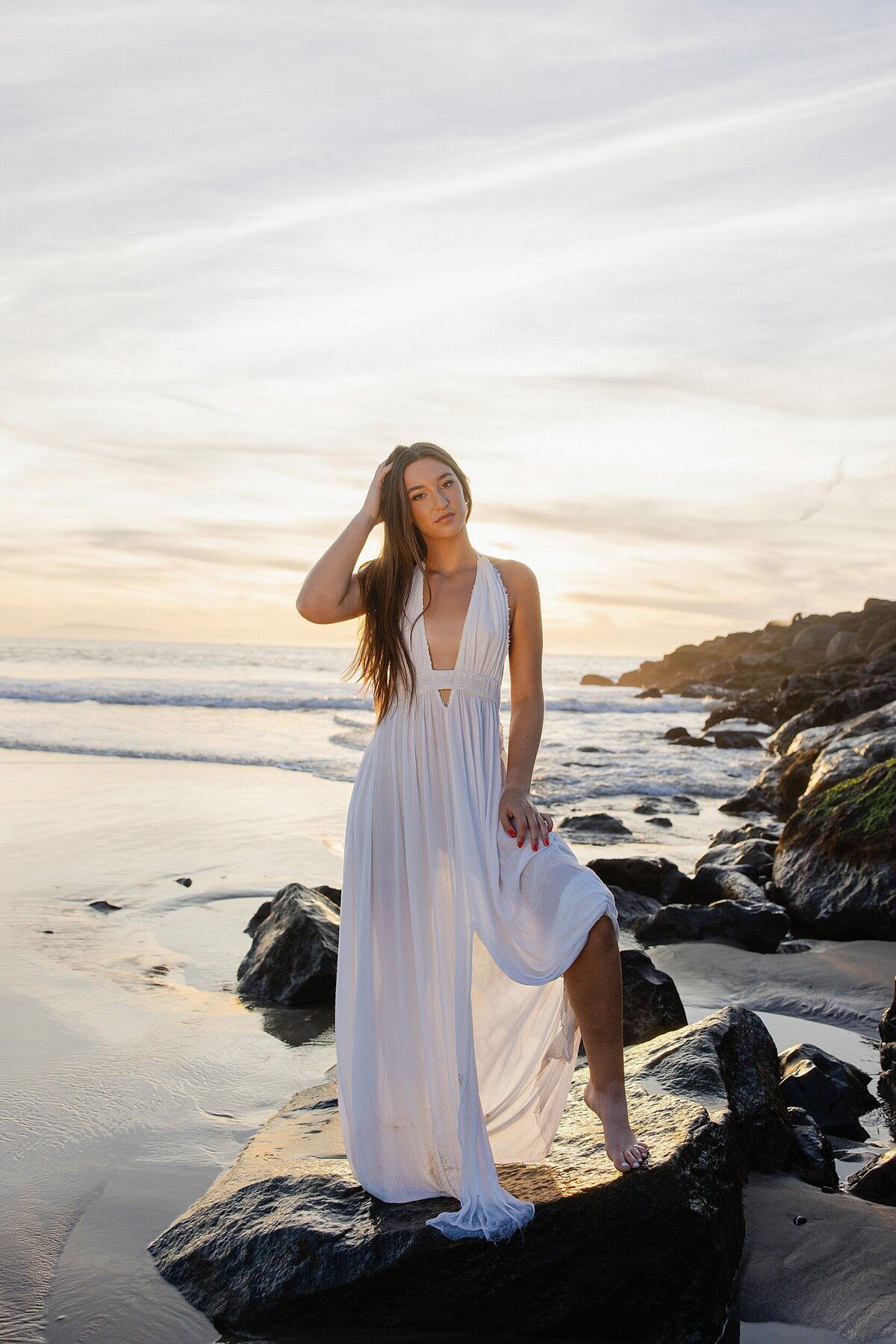 Dramatic sunset senior portrait of a teen girl standing in the sand with her hand in her hair, waves and sky glowing in the background, Ventura Beach