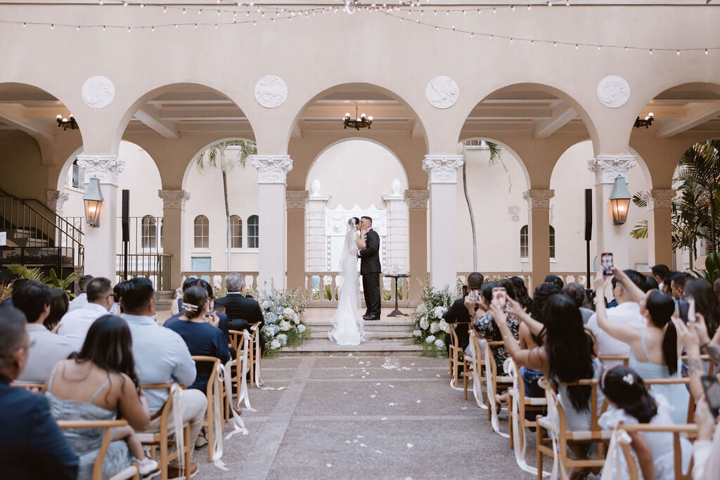 bride and groom kissing during ceremony under arches with guests watching in wishbone chairs