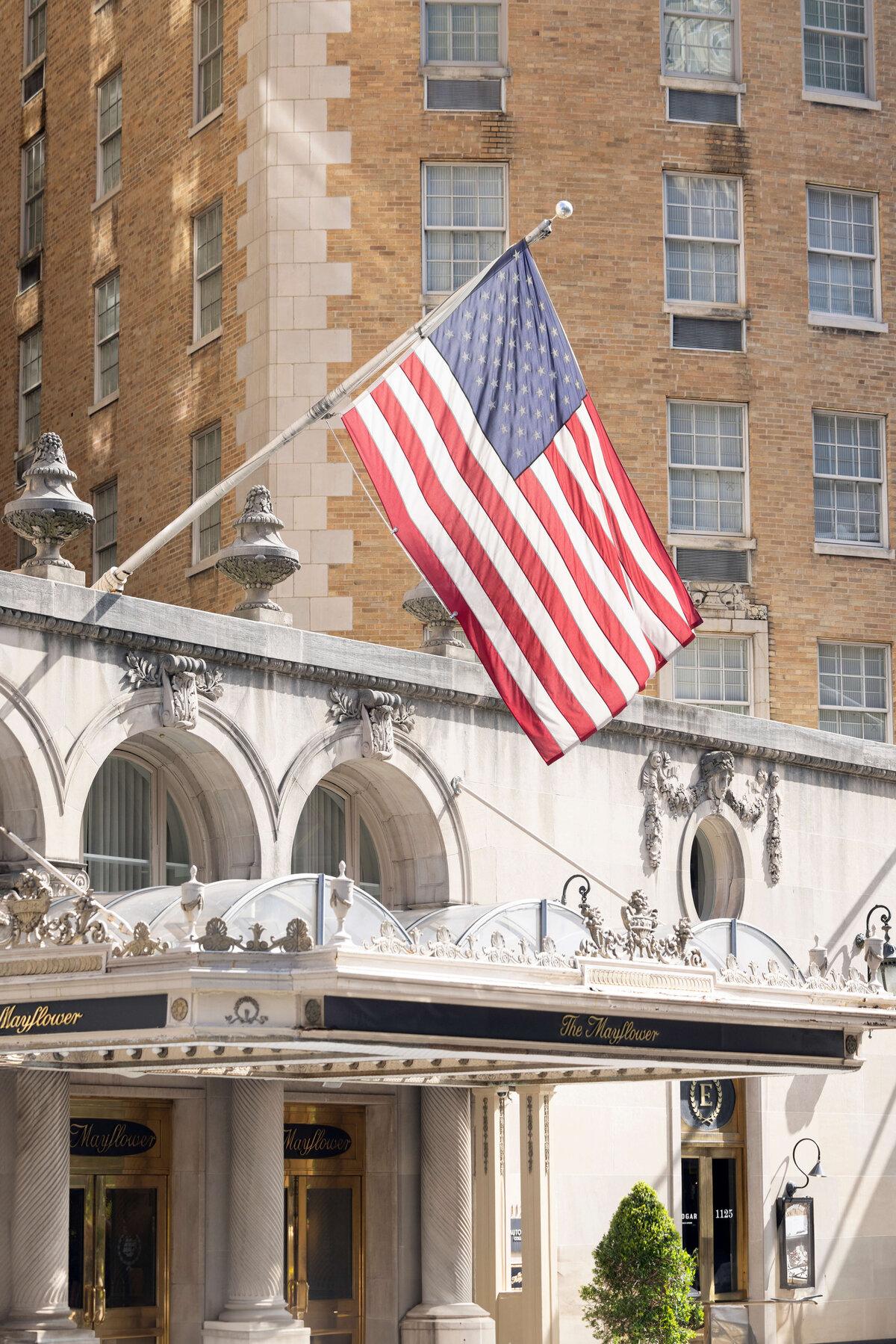 A large American flag waves in front of a historic building with ornate architectural details and tall windows. The building features a classic facade with columns and decorative arches under a clear sky.