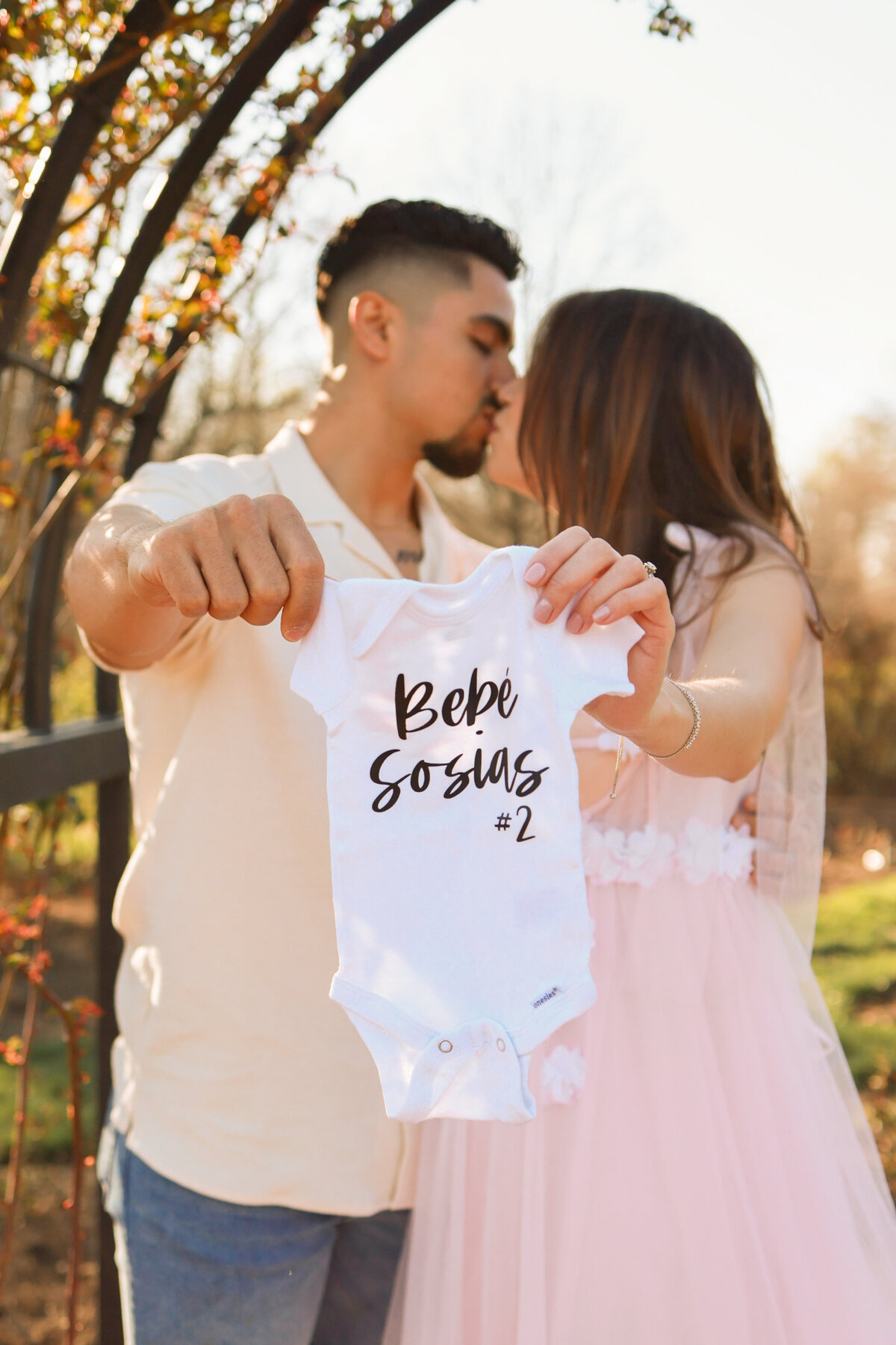 man and woman kissing while holding baby onesie saying Baby number two due