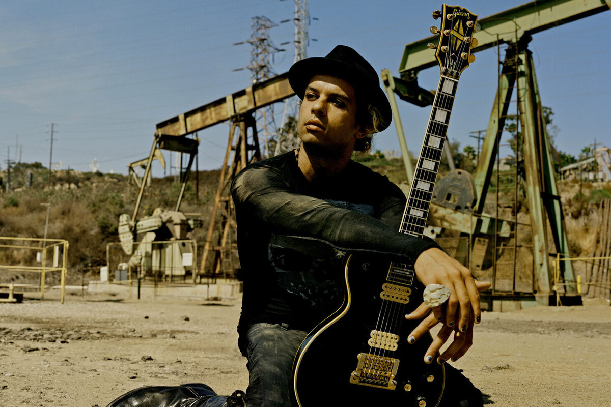 Musician Portrait Los Angeles Fernando Basillio sitting in sand in front of oil drill leaning on electric guitar upright in front of his knees Just Play Something Gallery