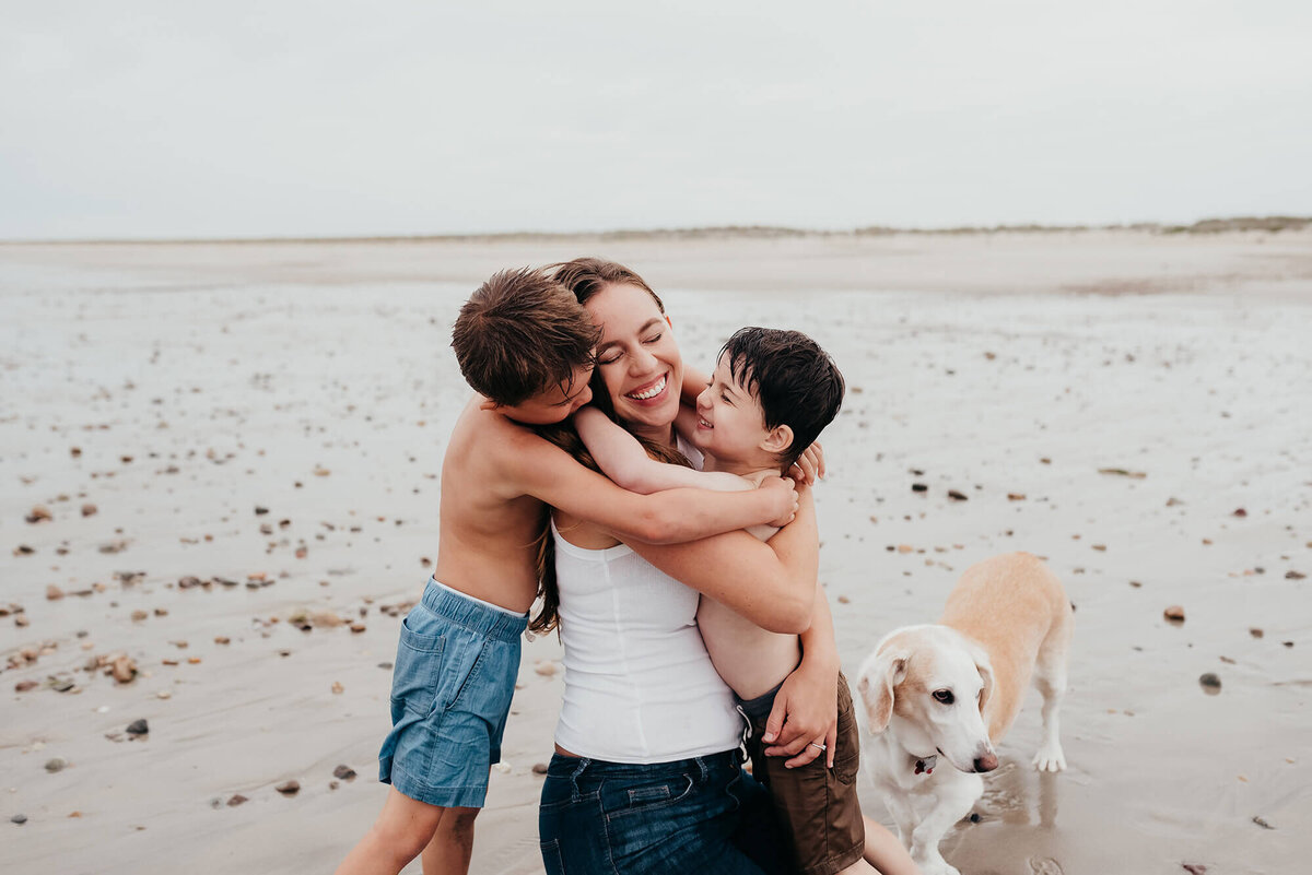 A mother kneels on the beach while being hugged by her two young sons, her senior dog  stands close by during their photoshoot with Allison Wolf, Boston Family Photographer