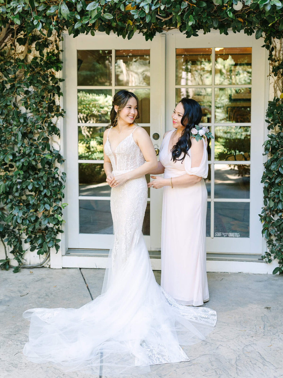 Bride in an elegant white lace gown and her mom in a pale pink dress smile at each other. They stand before glass doors.