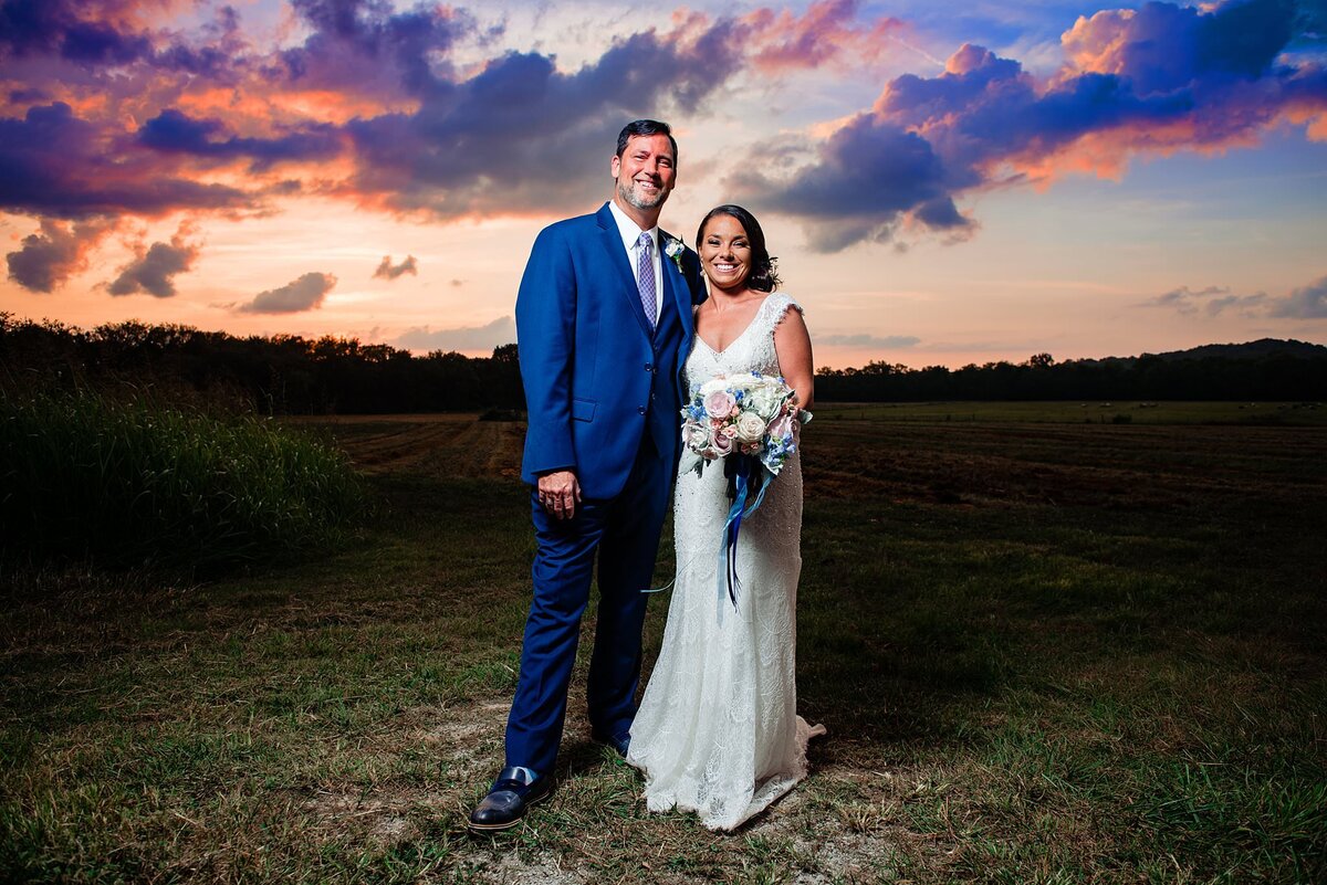 The bride and groom stand on top of a hill at Arrington Vineyards at sunset. The sky is painted with pinks, purples, blues and golds. The  groom is wearing a cobalt blue suit, white shirt and pink and blue pattered tie. The bride is wearing a white dress with a plunging neckline and lace detailing. The bridal bouquet is blush, burgundy and ivory with  cascading greenery and flowers.