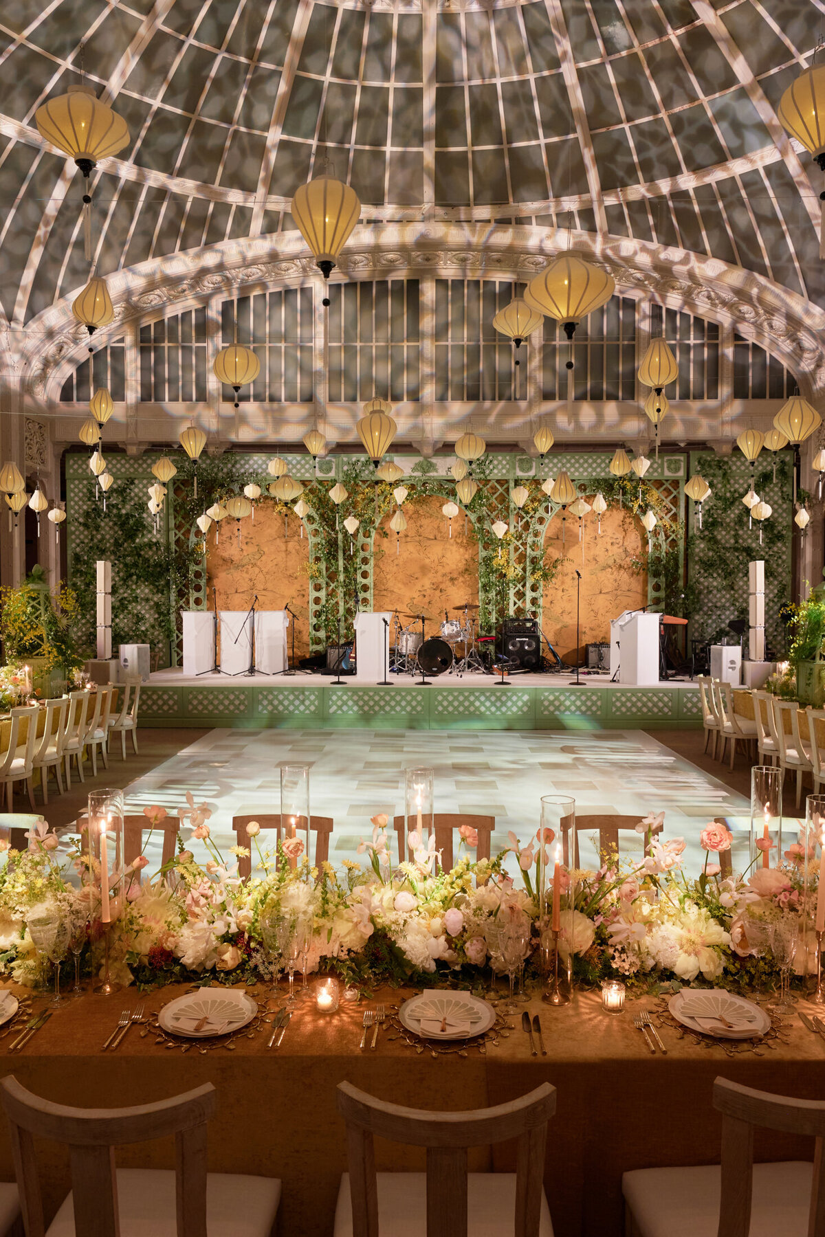 The elegant, glass-ceilinged venue, set up for a wedding reception, is located at The New York Public Library in New York City. Captured by photographer Shawn Connell, the scene showcases a beautifully decorated space with a dance floor surrounded by chairs and floral table arrangements. Lanterns hang overhead, adding a touch of charm to the setting. The stage, adorned with greenery and floral decor, is prepared for a musical performance, creating a romantic atmosphere for the event. Aaron Navak Films is also present as videographers, ensuring that every special moment is captured on video.