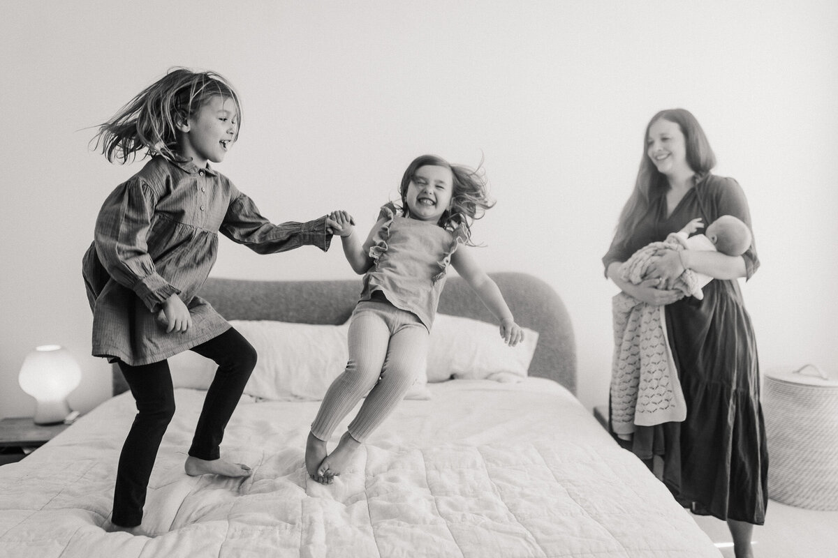 San Francisco sisters jump on bed while mom holds baby nearby at in home family photography session