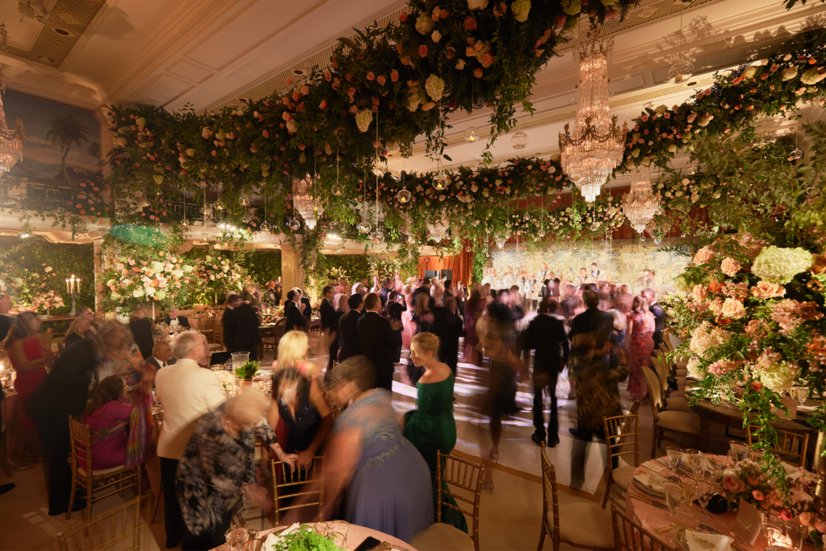 A vibrant wedding reception in a grand, elegantly decorated hall. Guests are dancing under lavish chandeliers and lush floral arrangements hanging from the ceiling. Tables are adorned with flowers and candles, and people are mingling and enjoying the festive atmosphere.