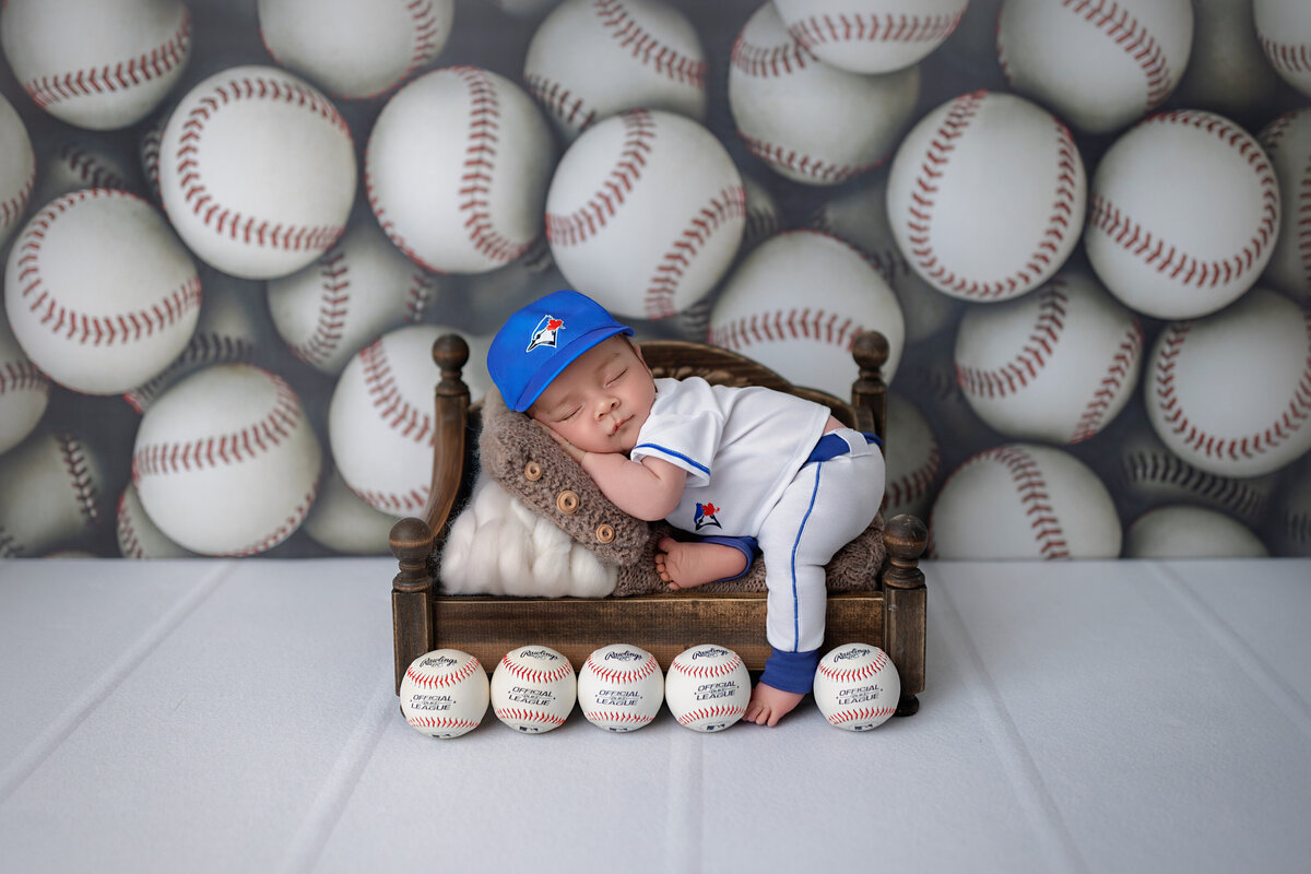 Newborn boy in a baseball-themed setup, sleeping on a mini bed with baseballs arranged in front and a sports backdrop behind him.