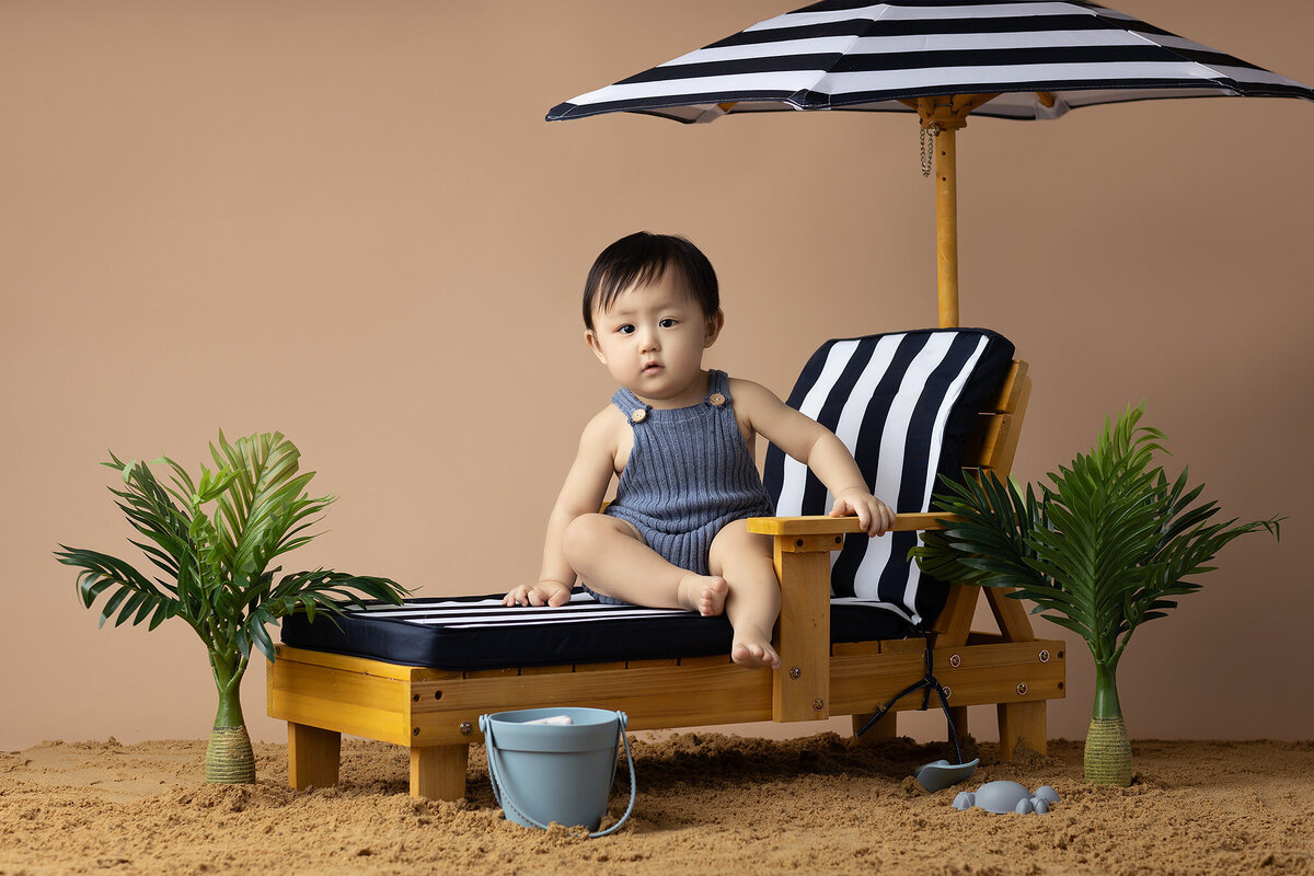 This adorable milestone photo captures a baby relaxing on a beach-inspired set complete with a striped umbrella, lounge chair, and tropical plants. The little one enjoys the summer vibes while holding a beach toy, creating a fun and playful image. The use of a beach theme in milestone photography helps preserve unique moments in your baby's first year. Dallas baby photographers bring creative setups like this to make your child's milestone session unforgettable. Trust Dallas baby milestone photographers to capture the essence of your baby's growth in a fun and artistic way.