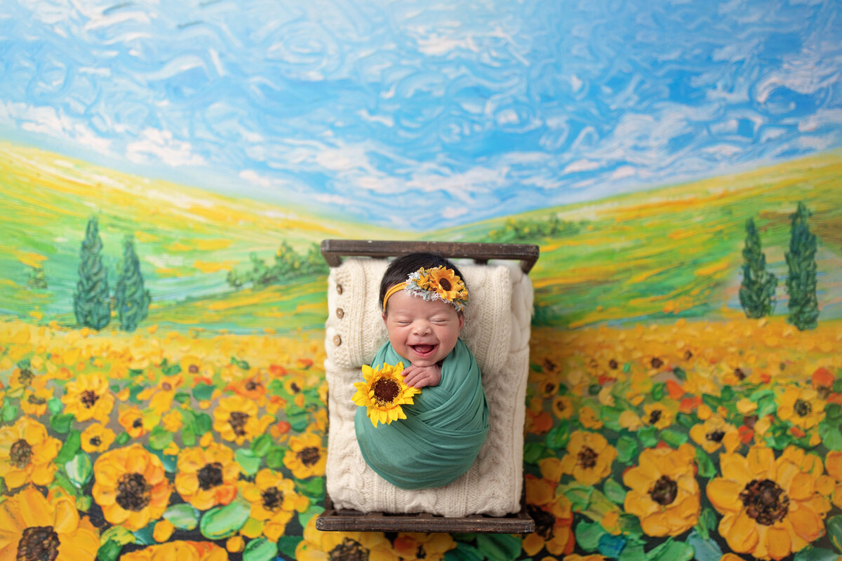 Newborn baby smiling in a sunflower field-themed setup with bright yellow flowers and a blue sky backdrop.