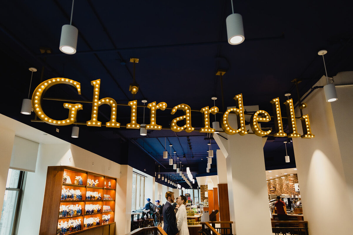 Just Married photo session couple stands under large lit Ghiradelli sign in Chicago store