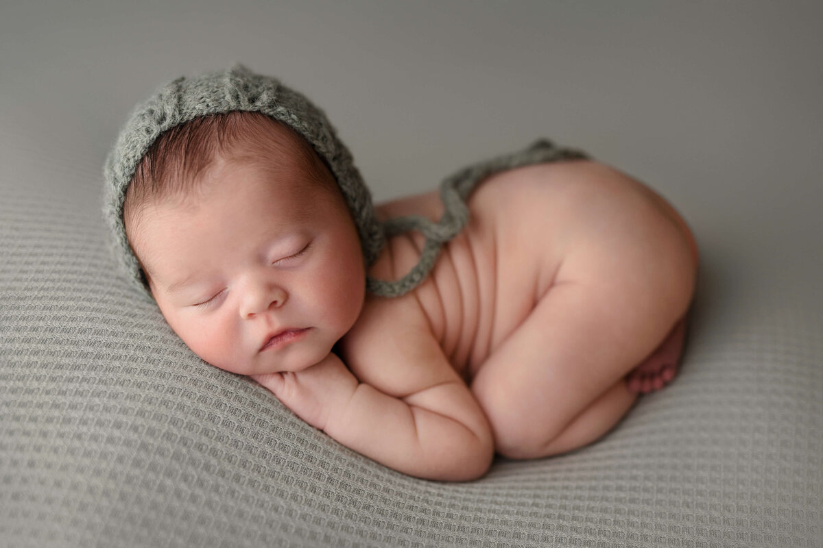 newborn boy scrunched up sleeping on his tummy on a green backdrop with green bonnet