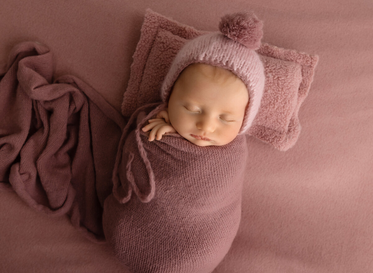 Brooklyn newborn photoshoot. Baby girl is wrapped in mauve wiht her fingers peeking out of the wrap. She is laying on mauve stretch fabric and a tiny mauve felt pillow. Captured by top Brooklyn newborn photographer Chaya Bornstein