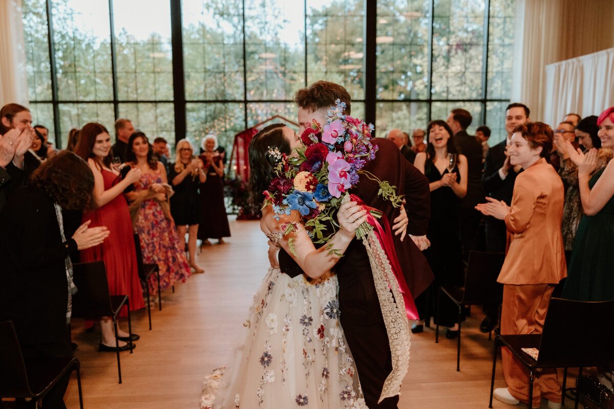 A bride and groom share a passionate kiss at the end of their wedding ceremony at the Roundhouse in Beacon, as guests applaud and celebrate around them. The bride holds a vibrant bouquet of colorful flowers, obscuring part of their embrace. She wears a floral-embroidered gown, and the groom is dressed in a dark suit. The large windows in the background reveal a scenic view of trees, adding to the festive and romantic atmosphere.