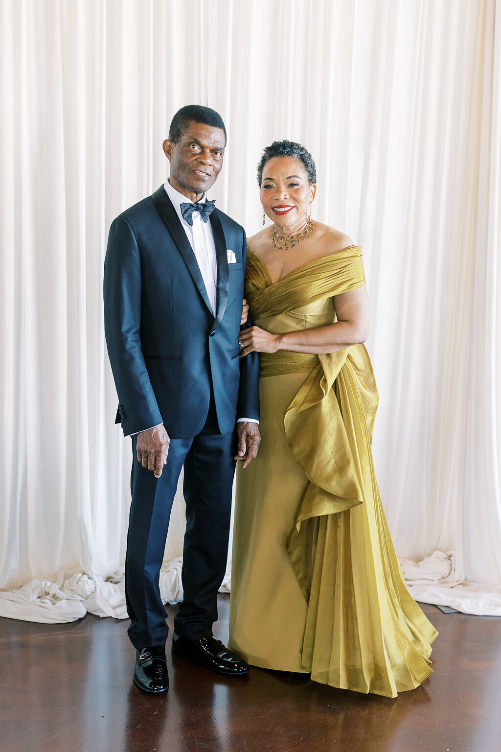 The groom’s parents stand together in a sophisticated gold and black ensemble, smiling warmly.
