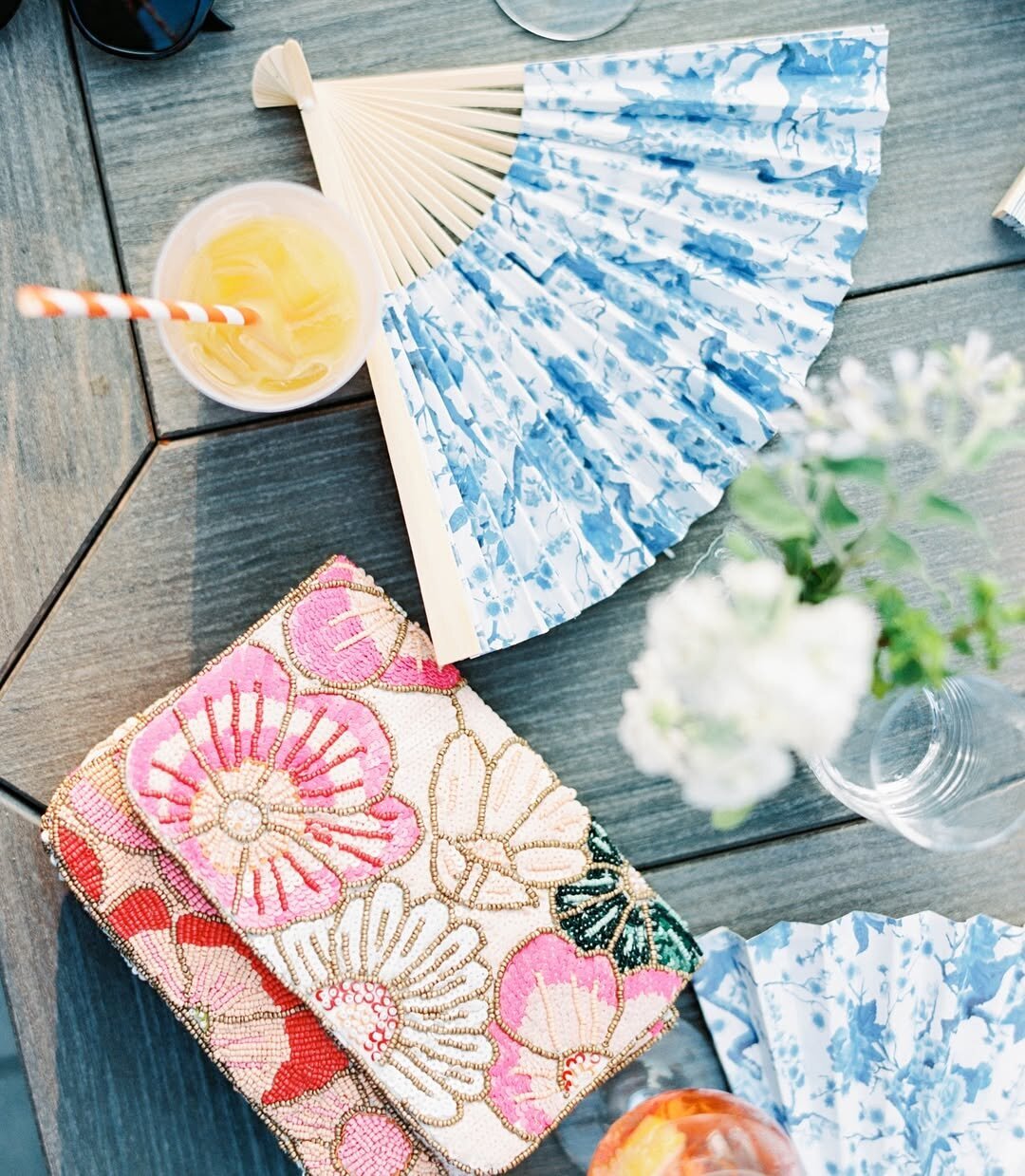 Wedding Photographer RI | A floral clutch, blue hand fan, and iced drink on a wooden table with a striped straw and blurred white flowers in a vase. Bright, summery ambiance.