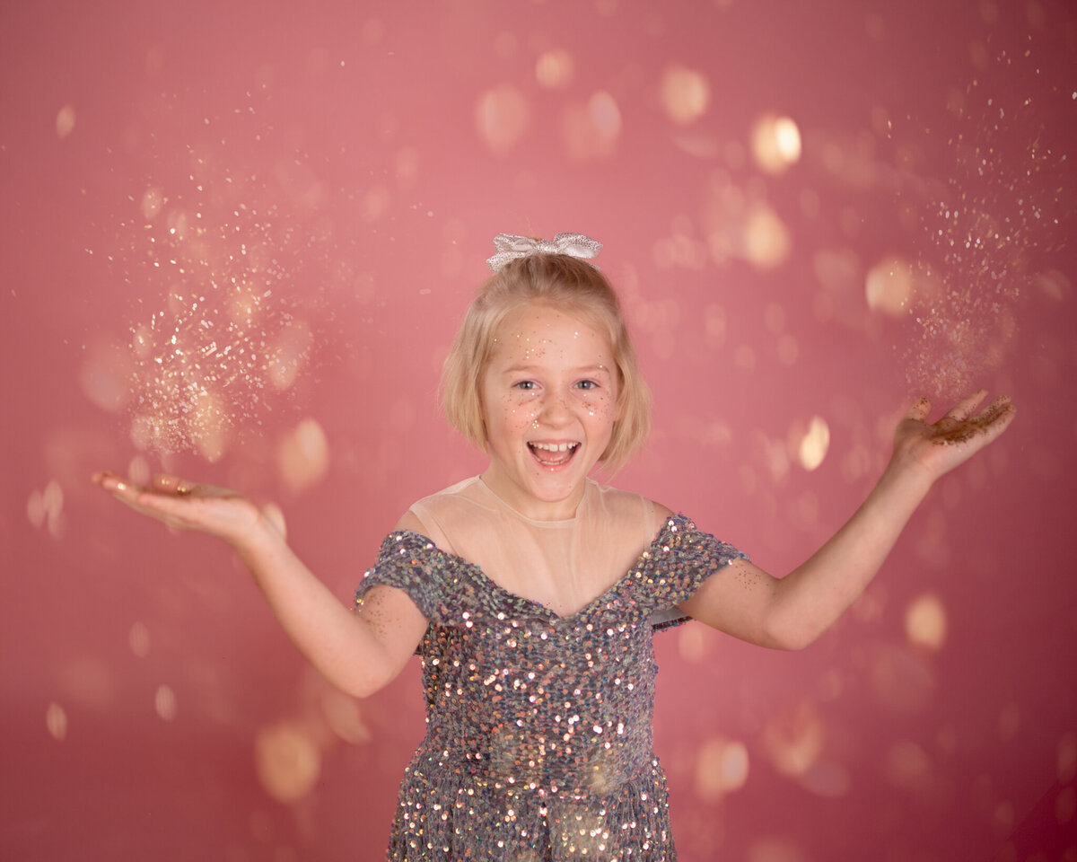 Whimsical portrait of a young girl surrounded by sparkling glitter against a pink backdrop, radiating joy and excitement. Captured in Overland Park, Kansas, this playful and vibrant setup is ideal for families seeking creative child photography in the Kansas City area.