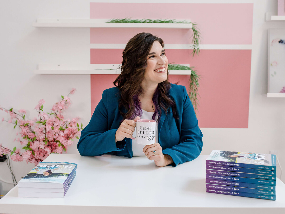 small business owner sitting behind a desk and holding a coffee mug with pink accents and her books piled next to her on the desk as she smiles for her branding photoshoot