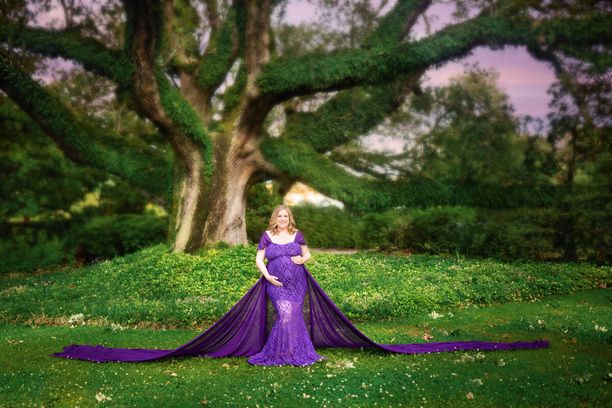 Pregnant woman with short blonde hair standing under a huge live oak tree in City Park.  She is wearing a purple lace mermaid gown from Mii-Estillo, with a long chiffon train.  The train is spread around her.  She has one hand on the top of her belly and the other on the bottom.  There is a purple sunset and mossy tree branches behind her.