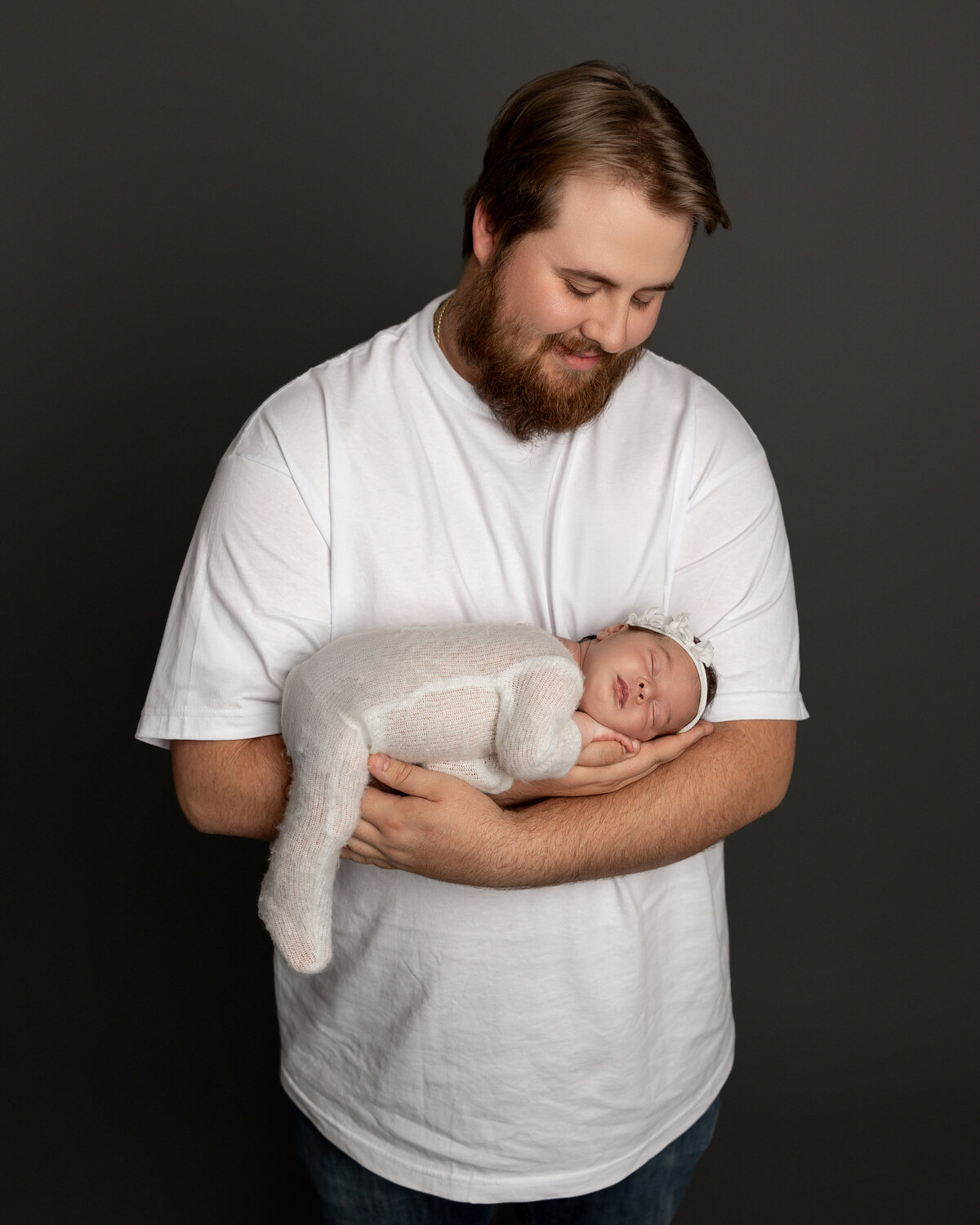Dad smiling down at his peacefully sleeping newborn daughter, swaddled in a soft cream knit outfit with a matching headband, cradled in his arms during their studio session. Captured by Inland Empire newborn photographer Kaitlyn Dawn Photography.
