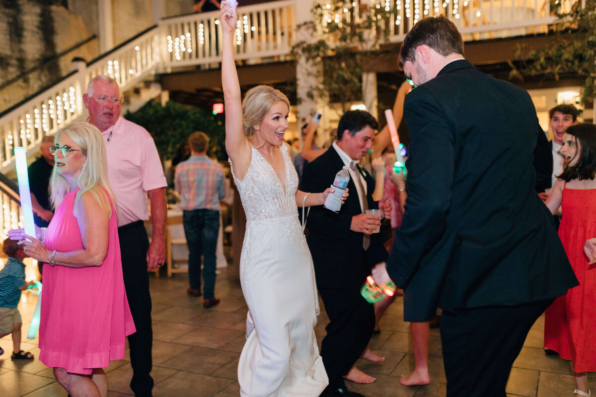 A bride in a white dress raises her arm joyfully while dancing with guests at a lively outdoor wedding reception, surrounded by people in colorful attire and festive lights.