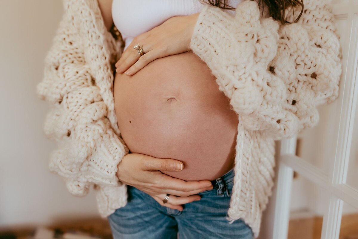 Person wearing a knit sweater and jeans, holding their pregnant belly with both hands.