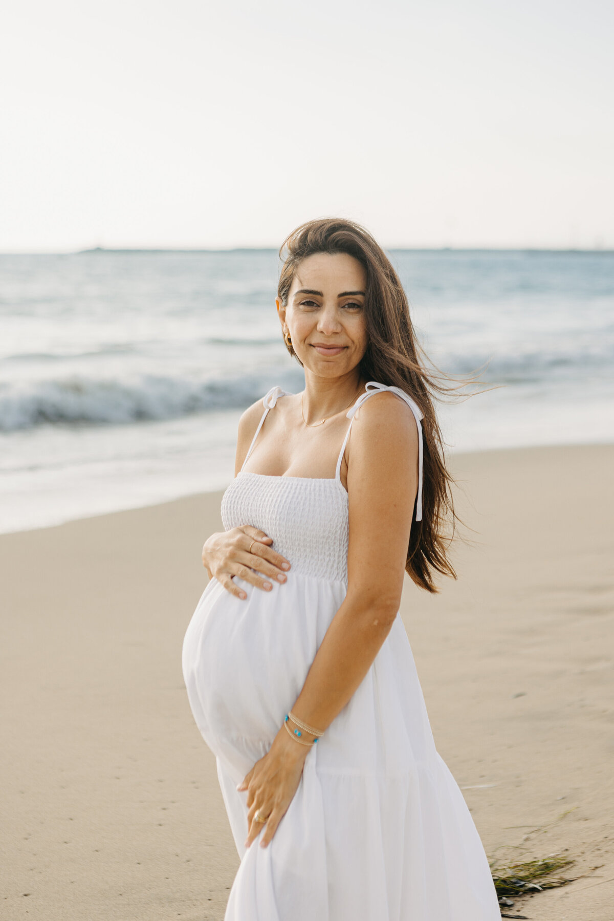 Pregnant woman in white dress cradles bump on the beach in Los Angeles.