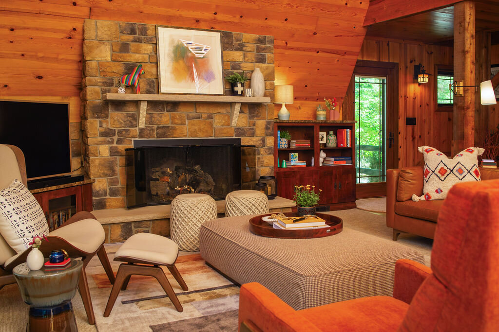 A warmly lit wood cabin living room featuring a prominent stone fireplace with a wooden mantel adorned with artwork and decorative objects. An orange armchair and a tan armchair with a patterned pillow are positioned near a patterned ottoman serving as a coffee table. A brown leather sofa with a patterned pillow is partially visible on the right. A flat-screen TV sits to the left of the fireplace.