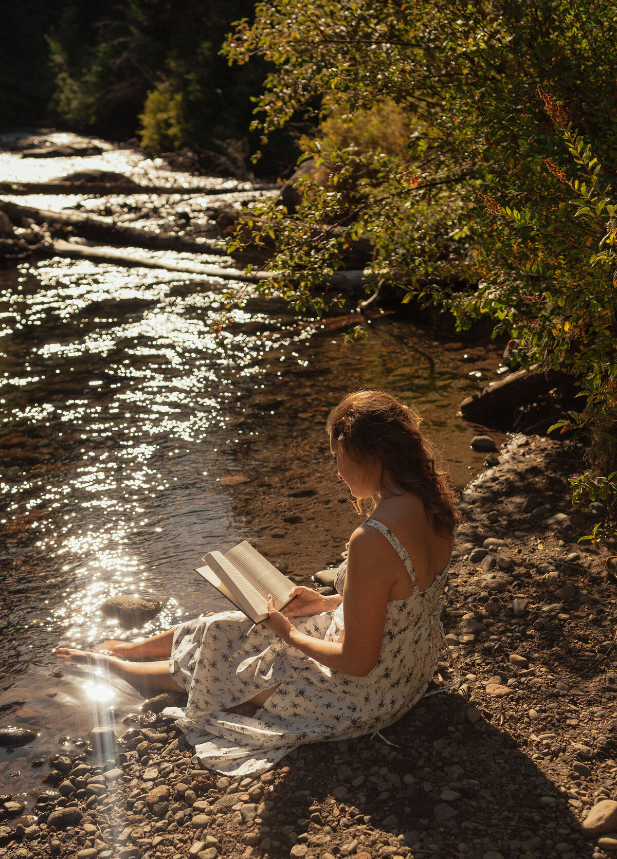 woman in white dress sits next to the river and reads a book while her toes dip in the water