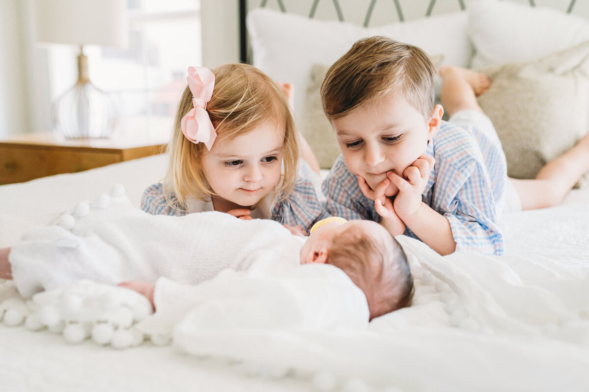 young brother and sister inspect brand new baby while lying on bed