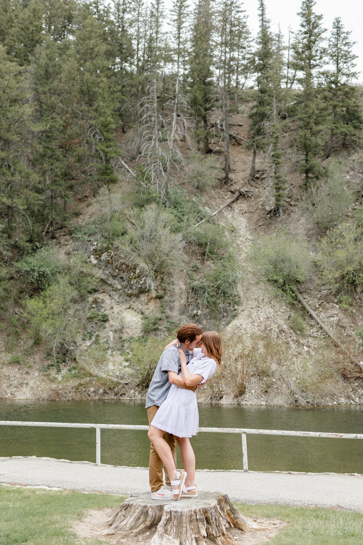 A couple stands on a tree stump by the river, sharing a kiss with the towering forest in the background at Vivian Park, Provo Canyon.