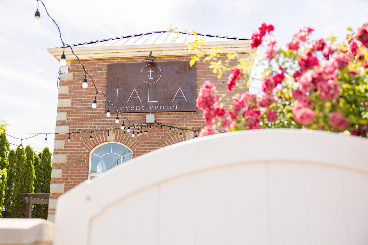 A beautifully decorated brick building with the "Talia Event Center" sign, adorned with string lights and vibrant pink flowers.