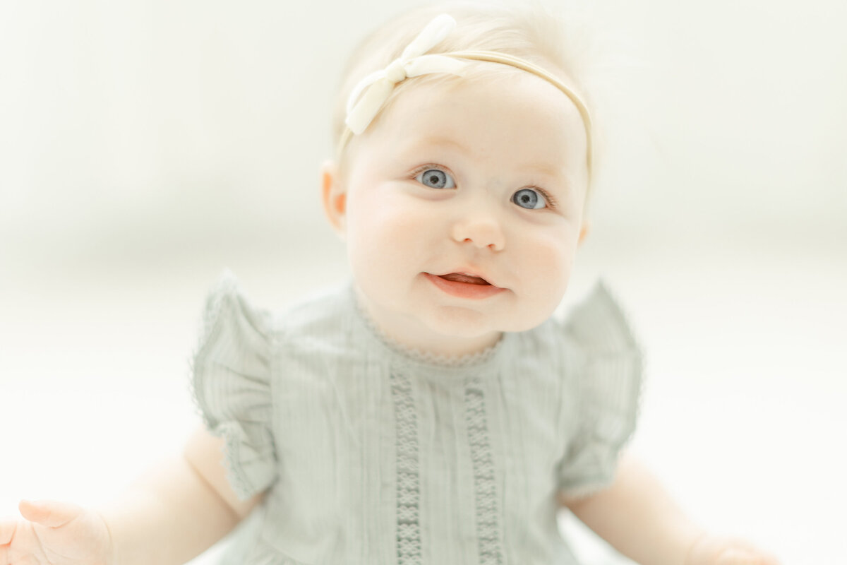 A blue eyed baby girl sitting on the floor in a photography studio for her milestone photos.