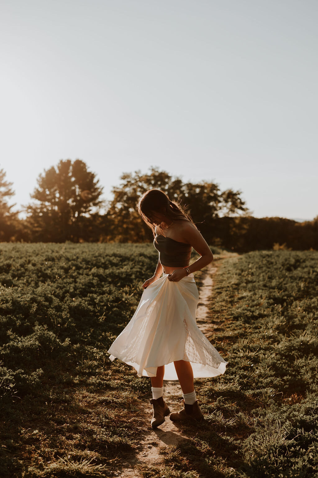 A woman in a sleeveless top and flowing white skirt stands on a dirt path in a sunlit field, surrounded by greenery and trees. Captured by a New England Portrait Photographer, the scene is bathed in warm sunlight, creating long shadows and a serene atmosphere.