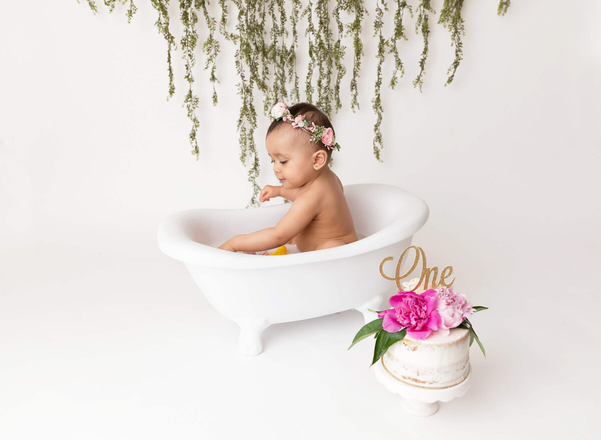 Baby girl sits in a bathtub and plays with water during a first birthday cake smash.