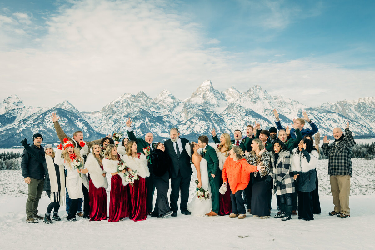 A bridal party at Glacier View overlook after a Jackson Hole Wyoming wedding in the winter snow celebrating the new bride and groom kissing