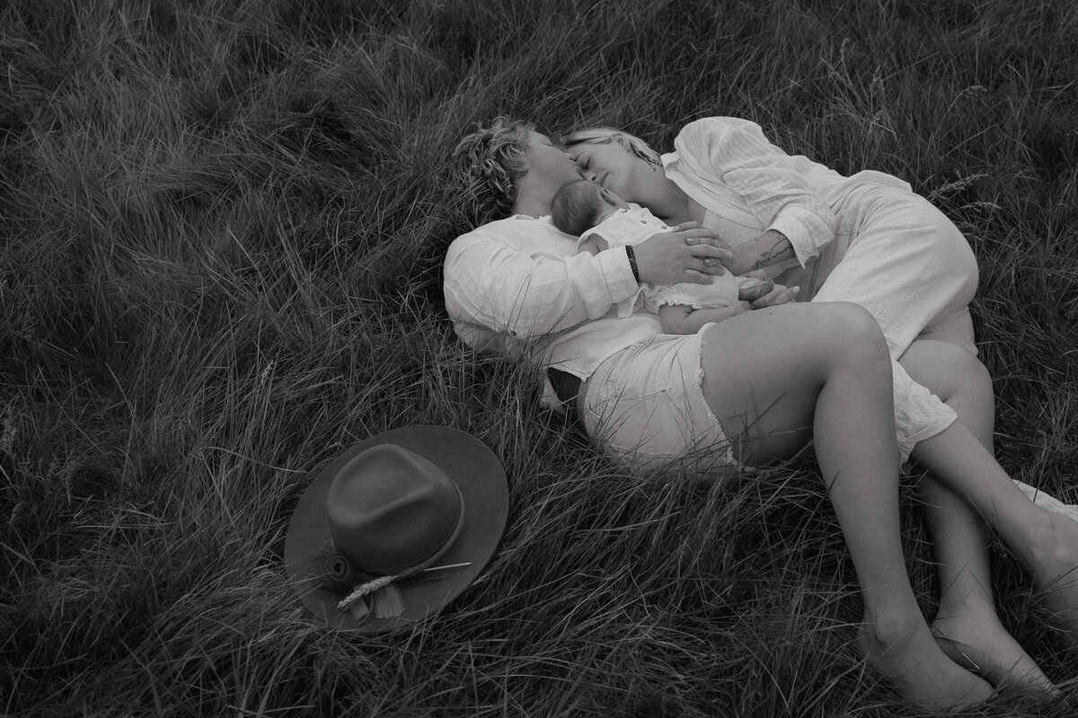 A black-and-white image of a couple lying in a grassy field with their newborn, reflecting a luxury and artistic wedding photography style.